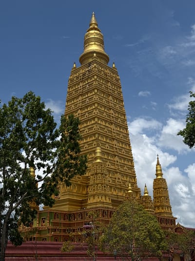 A tall, golden Buddhist stupa stands against a bright blue sky with scattered white clouds. The stupa is intricately decorated with rows of small niches and geometric patterns. Several smaller golden spires surround the base of the main stupa. In the foreground, lush green trees with dark trunks partially obscure the lower sections of the structure. The base of the stupa is adorned with green and red accents. The overall scene depicts a religious architectural landmark in Na Nuea, Thailand, under a sunny, partly cloudy sky.