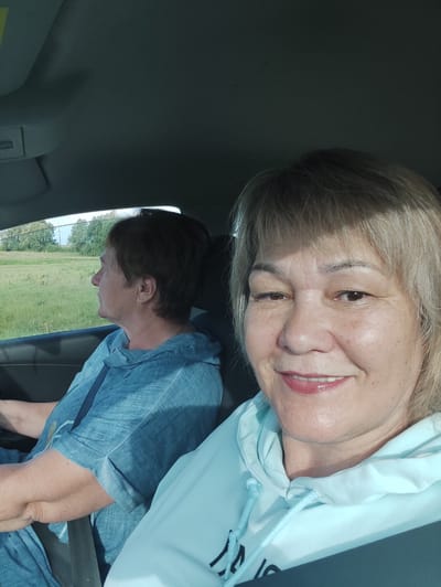 Two women are in the front seats of a car, with one woman in the foreground taking a selfie. The woman in the foreground has blonde hair and is smiling, wearing a light blue hoodie. She has makeup on and appears to be in good spirits. The other woman is in the driver's seat, looking out the window to her left. She has short brown hair and is wearing a blue shirt. The car appears to be moving through a rural area, with green fields and trees visible outside the windows. The sun is shining, suggesting it is daytime. The overall scene suggests a casual road trip or outing. No visible text is present in the image.