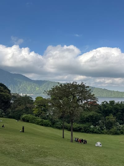 The image captures a scenic view in Baturiti, Indonesia. The foreground is dominated by a lush green grassy field, which slopes gently downwards. Several people are scattered across the field. In the mid-ground, a group of about eight individuals is gathered, appearing to be engaged in a conversation or activity. A small, white, dome-shaped structure, possibly a tent or a shelter, is visible further up the slope, to the right of the group. A lone figure is seen walking on the field in the lower right portion of the image, and another person is visible further in the distance. Two tall trees with dark bark and full canopies stand prominently in the middle ground, framing the scene.
The background features a dense, verdant forest that climbs up the side of a large mountain. Beyond the mountain, a body of water, likely a lake, is visible, with hints of buildings or settlements along its shores. The sky is a vibrant blue, partially obscured by large, dramatic white clouds that add depth and texture to the scene. The lighting suggests it is daytime, likely late morning or early afternoon, with clear visibility. The overall impression is one of a peaceful, natural landscape.