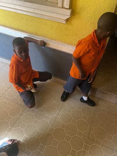 A young boy is sitting on a tiled floor in an unknown location in Markurdi, Nigeria. He is wearing an orange shirt and grey shorts, and black shoes with white socks. The wall in the background is painted yellow and blue. Another boy is sitting on the floor close to the first one. He is also wearing the same uniform, including orange shirt, grey shorts, and black shoes with white socks. The floor is tiled in a light brown geometric pattern.