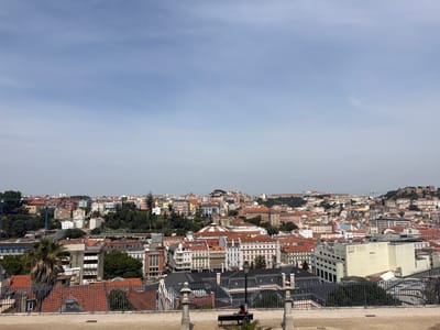 An elevated, wide-angle, eye-level view captures a sprawling cityscape of Lisbon, Portugal, under a hazy, pale blue sky. The foreground features a tan, textured surface, possibly stone or concrete, leading to a dark metal fence. Beyond the fence, a black bench with a person sitting on it is visible, facing away from the camera. Two stone busts on pedestals are positioned on either side of the bench. The midground and background are filled with a dense collection of buildings, characterized by red-tiled roofs, white and pastel-colored facades, and varying architectural styles. Numerous trees are interspersed throughout the urban landscape. In the distance, a prominent hilltop structure, likely a castle or fortification, overlooks the city. The overall scene depicts a sunny day in a densely populated urban environment.