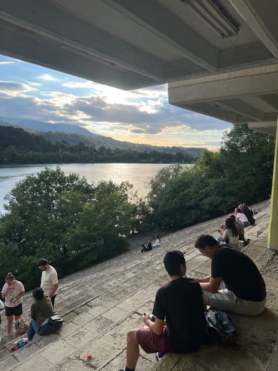A group of people are gathered on a tiered concrete structure overlooking a wide body of water, likely Pancharevo Lake in Pancharevo, Bulgaria. The scene appears to be set during the late afternoon or early evening, with the sun casting a warm glow across the sky and reflecting on the water. The sky is partly cloudy, with rays of sunlight breaking through, illuminating the distant hills and mountains that surround the lake. Lush green trees line the shore, creating a natural backdrop to the urban infrastructure.
Several individuals are seated on the concrete steps, some engaged in conversation, others looking out at the view. In the foreground, two young men are sitting together, one holding a phone. Further up the steps, a man and a woman are standing and interacting, while other groups are scattered across the tiered seating area. The concrete structure itself appears to be part of a larger building or amphitheater, with a concrete overhang providing shade in the upper portion of the image. The overall atmosphere suggests a relaxed gathering or a scenic overlook point for locals and visitors to enjoy the natural beauty of the area.