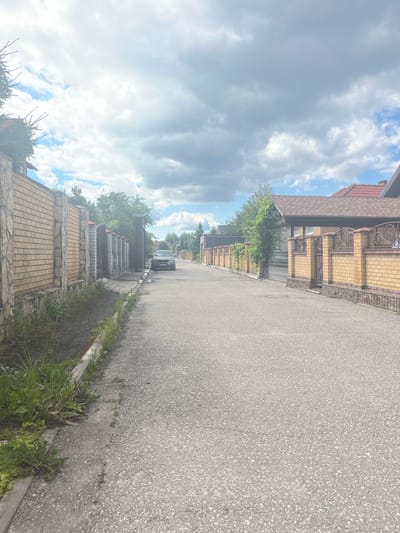 A street in Orekhovka, Russia, is shown from a street-level perspective. The asphalt road occupies the majority of the foreground and midground, with grassy areas and weeds growing along the sides. Brick fences with stone accents and decorative metalwork line both sides of the street, separating properties. Houses with tiled roofs are visible behind the fences. A dark-colored car is parked on the side of the road in the midground, and trees and foliage are present throughout the scene. The sky above is partly cloudy with a mix of gray clouds and patches of blue sky.