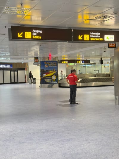 The image depicts a clean, well-lit airport terminal, likely an arrivals or baggage claim area, that appears relatively quiet. In the central foreground, a person wearing a red shirt with "Jet2.com" emblazoned on it stands, facing right and looking at a mobile device. This individual is presumably an airport or airline staff member. Behind them, empty baggage carousels are visible, suggesting a period of low activity.
To the left, prominent overhead signs guide passengers. One sign displays "Information Information Información" (Information in English and Spanish) with an 'i' symbol and an arrow pointing left. Above it, the "aena" logo confirms the location as a Spanish airport. Below this, another sign reads "Ausgang Exit Salida" (Exit in German, English, and Spanish), accompanied by a door icon and an arrow also pointing left.
Further in the background, another traveler with luggage can be seen walking away. Against a pillar, a large blue and yellow advertisement for "Submarine Safaris Amarilla Marina" is displayed, featuring a submarine and text promoting "The Underwater Adventure of a Lifetime! BOOK HERE" along with a QR code and website (www.submarinesafaris.com). Near the background on the left, a "WHSmith" store is also visible.
The floor is a textured light blue-grey, complementing the white grid ceiling which houses various lighting fixtures and circular vents. The visible multilingual signage (German, English, Spanish) on the exit and information signs, coupled with the "aena" logo and "Jet2.com" branding, indicates an international airport setting, consistent with the location context of Granadilla de Abona, Spain. The scene conveys a calm, organized daytime atmosphere.
