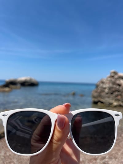 A close-up shot shows a pair of white-framed sunglasses with dark lenses held up by a person's hand. The hand has neatly manicured nails with a light pink polish. The background is blurred, but it is evident that the scene is a beach in Sutomore, Montenegro. The sea is visible, with calm, clear blue water. In the distance, there are large, rocky formations jutting out of the water. The sky is a clear, bright blue, suggesting a sunny day. The overall impression is one of a relaxed summer day by the sea.