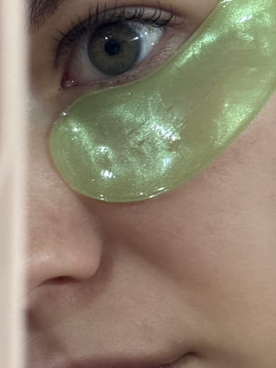 A close-up, indoor shot shows a person's eye with a green, shimmering under-eye mask applied. The person's eye is open, and the mask partially covers the lower portion of the eye and the skin beneath. The nose and part of the other eye, which is closed, are also visible. The person is lying on something light-colored, possibly a pillow or a surface. The location is not determinable from the visual information provided.