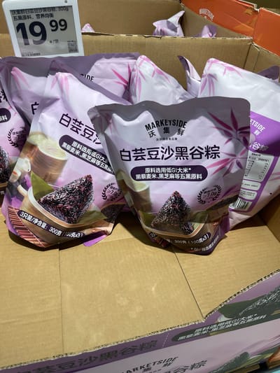 The image shows a close-up view of packaged food items in a cardboard box. The packaging is predominantly purple and white with Chinese characters and images of food. The items appear to be a type of rice dumpling, possibly zongzi, filled with black grains and possibly other ingredients.
In the foreground, a package is prominently displayed, featuring a dark, triangular-shaped food item nestled in a wooden bowl with green leaves. The text on the packaging indicates it is "Marketside" brand, with Chinese text that translates to "Black Rice and Five-Grain Zongzi" and mentions "Low GI ingredients." There's also nutritional information visible on some of the packaging.
A price tag is attached to the front of the box, showing "19.99" with Chinese characters, suggesting a price in Yuan. The background shows more of the same packaged food items within cardboard boxes. The overall environment appears to be a retail setting, possibly a grocery store or market. The location is identified as Panyu District, China.