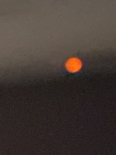 The image is a low-resolution, grainy photograph of an out-of-focus orange circle, possibly the sun or a light source, set against a dark, indistinct background. The background appears to be a gradient of muted grays and dark browns, suggesting a sky at dusk or dawn, or possibly a smoky or hazy atmosphere. There is no identifiable terrain, structures, or individuals in the image. The location is given as Stallings, United States of America, but the image itself provides no visual cues to confirm this.