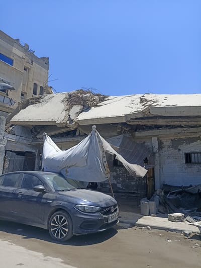 This is a photograph taken from street level, looking towards a damaged building in the city of Gaza, Palestinian Territories. The building's roof is heavily damaged, with concrete slabs broken and debris scattered. Exposed rebar is visible. A white tarpaulin, torn and hanging, is supported by poles in front of the damaged structure. To the left of the building, another partially damaged building can be seen, with a blue-tinted window visible. In the foreground, a dark blue sedan is parked on the dirt and asphalt road. The car has a license plate that reads "3-3598-06". The environment appears to be an urban setting, with evidence of destruction and debris on the ground. The sky is clear and blue.