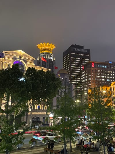 Here is a description of the image.
This is a night cityscape of Shanghai, China. In the foreground, there is a street lined with trees and benches occupied by pedestrians. There are several older, grand architectural buildings illuminated with yellow light, including one with a prominent golden crown-like structure. In the background, there are modern high-rise buildings, some illuminated with neon signs. The overall atmosphere is vibrant, with a mix of historical and contemporary architecture.