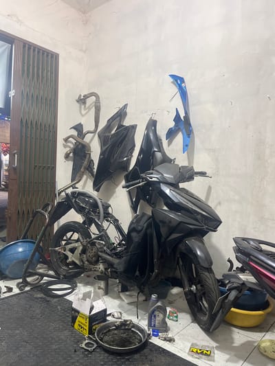 An indoor, dimly lit motorcycle repair shop in Ciakar, Indonesia, features a disassembled black motorcycle as the central subject. The bike's frame and engine are exposed, with various parts scattered around. To the left, a blue basin and a metal stand are visible. The wall behind the motorcycle is adorned with several detached black and blue plastic fairings and a metal exhaust pipe. On the floor, there's a roll of black material, a metal pan containing dark fluid, a box labeled "RVN CYLINDER KIT," a bottle of "FUKUOKA OIL," and other miscellaneous items. A metal accordion gate is partially visible on the far left. No people are present in the scene.