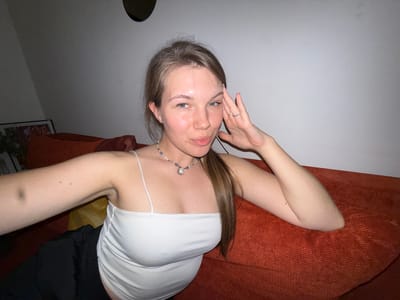 A young woman is lying on an orange textured surface, likely a couch or bed, in an interior setting. She is wearing a white spaghetti-strap top. She has a necklace, and a ring on her left hand. One arm is raised and her fingers are near her face. The background includes a white wall and what appears to be a black garment.
There is no information in the image that indicates the exact location is in the city of Vozdovac Urban Municipality, Serbia. The image does not provide enough forensic information.