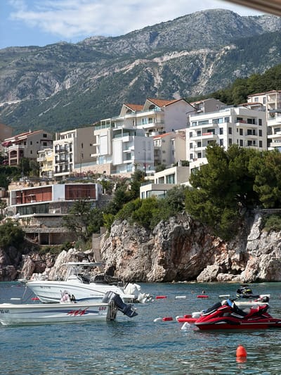 In Pržno, Montenegro, a coastal town nestled below a mountainous backdrop, the scene unfolds along a rocky shoreline dotted with modern white buildings. The azure water in the foreground is populated by various watercraft, including a white speedboat labeled "Car Camarat," and a smaller white boat with "BD 441" visible on its hull. A red jet ski, with a person seated on it, is positioned towards the right of the frame. Several buoys, both red and white, are scattered across the water's surface, marking designated areas. Behind the boats and the rocky cliffs, multi-story buildings with white facades and prominent balconies climb the hillside, interspersed with lush green trees. The imposing mountain range, characterized by its rugged, stony slopes and scattered vegetation, dominates the upper portion of the image, with a clear, partly cloudy sky above.