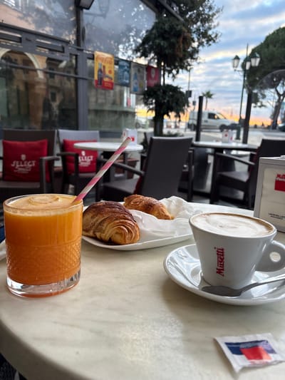 A table displays a delightful breakfast scene. A cup of cappuccino sits elegantly on a saucer with a spoon, alongside a small package of sugar. A delicate white dish holds two golden croissants. In the foreground, a glass of orange juice with a pink-striped straw completes the meal. The table overlooks an outdoor setting featuring trees, streetlights, and building facades. The view suggests a relaxed cafe ambiance.