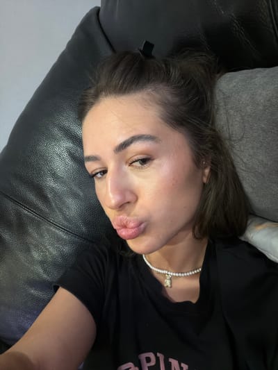 A woman is captured in a close-up, selfie-style photograph, lying down and looking directly at the camera with her lips puckered. She is wearing a black t-shirt and a pearl necklace with a small, gold-colored bear charm. The background consists of a dark, textured surface, likely upholstery, and a gray cushion. The lighting suggests an indoor setting, possibly a living room or bedroom. There are no identifiable landmarks or environmental cues that would specifically place the scene in Novi Iskar, Bulgaria.