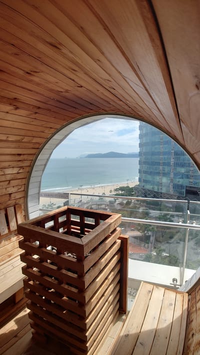 This is a medium shot of a sauna room with a view of Haeundae Beach in Busan, South Korea. The sauna is constructed of wood and features a large, circular window that overlooks the beach, the ocean, and a modern skyscraper. Inside the sauna, a wooden bench and a wooden heater are visible. The beach is populated with many people, and there are trees and a walkway between the beach and the building. The sky is cloudy.