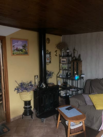 This image depicts an indoor scene, likely a living room, in Sabadell, Spain. The room features a black wood-burning stove on the left, with a tall black chimney pipe. In front of the stove, a woven basket filled with dried purple flowers sits on a black ornate stand. To the left of the stove, a framed landscape painting hangs on a yellow wall, depicting a house, trees, and a cloudy sky. Above the stove, a small decorative sunburst hangs on the wall. To the right of the stove, a black metal shelving unit holds various decorative items including vases, framed photos, and a small wooden cabinet. An essential oil diffuser with a blue and green light is visible on one of the shelves. Further to the right, a brown sofa with throw pillows, one yellow and one purple, is partially visible. In the foreground, a light-colored wooden coffee table with a woven basket underneath holds a remote control and a small metallic object. The ceiling is made of dark wood planks, and the walls are a mixture of yellow and textured gray paneling. The floor is tiled with terracotta-colored squares. The overall impression is a cozy, somewhat rustic interior.