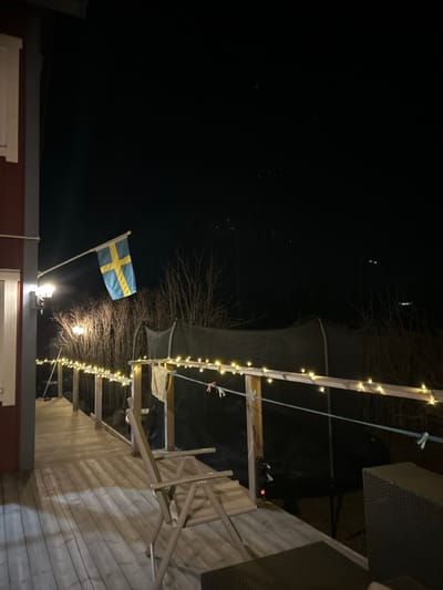 The image depicts an outdoor residential area at night in Gränsta, Sweden, characterized by artificial illumination against a dark, starry sky. On the left, a portion of a red wooden building with white trim is visible, from which a wall-mounted light fixture extends. A Swedish flag, blue with a yellow Nordic cross, is affixed to a pole extending from this building, positioned to the left of the frame's vertical center.
A wooden deck extends from the building into the scene, featuring a railing composed of vertical posts and a horizontal top rail. A string of warm-white LED lights is draped along the entirety of the visible top rail. On the deck, a light-colored, foldable outdoor chair is present, facing away from the viewer. In the bottom right corner, a dark, woven outdoor furniture piece is partially visible.
A light-colored cloth item is draped over the deck railing, and a blue rope is strung horizontally below the top rail, adorned with several multi-colored clothespins. Beyond the deck and railing, the background consists of a dark environment. Bare deciduous trees or bushes are discernible in the mid-ground. Behind these, a large, dark mesh enclosure, consistent with a trampoline safety net, is visible. Faint light sources are present in the distant background. No individuals or active interactions are depicted in the scene.