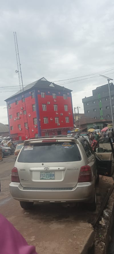 The image captures a bustling street scene in Umuahia, Nigeria, under an overcast sky. A silver Toyota Highlander with an open door is the foreground subject, its license plate reading "FST-218BM." Stickers adorn the rear window. The backdrop features a five-story building painted red with a blue top, branded with "R&S" logos. Adjacent to it is a gray building. A tall communications tower stands behind the red building. Numerous people, vehicles, and market stalls fill the street, indicating a busy urban area.