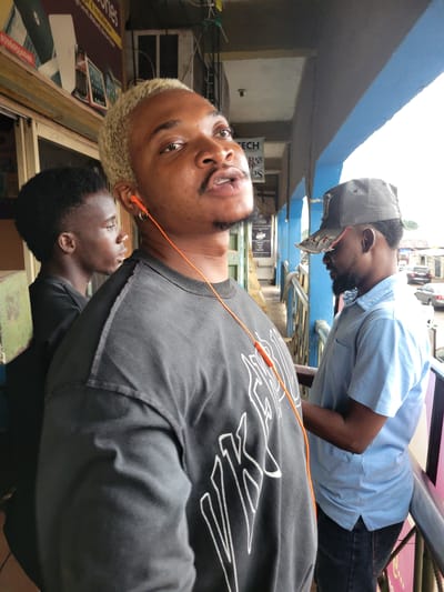 The photo captures three young men in what appears to be a commercial area of Makurdi, Nigeria. One man, facing the camera, has bleached blonde hair and is wearing a black shirt with white graphic designs and orange earphones. Another man stands behind him, wearing a black shirt. A third man, partially visible in the background, wears a light blue button-down shirt and a grey baseball cap. The setting includes the facade of shops and buildings, hinting at an urban commercial district. A sign for "TECH" can be seen in the background.