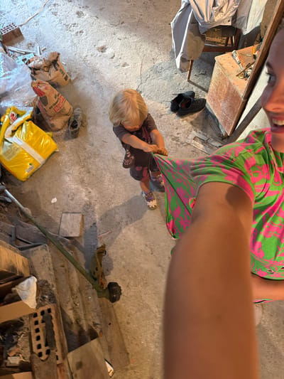 An overhead, high-angle, medium shot captures an indoor scene with a concrete floor and dimly lit surroundings. A young child with blonde hair, wearing a dark t-shirt and shorts, stands in the center of the frame, holding onto a green and pink patterned fabric. The fabric appears to be part of a garment being pulled by the child. To the right of the child, an adult's arm and torso are visible, also in a green and pink patterned shirt, suggesting a selfie-style capture. The background features several objects, including bags of what appear to be construction materials, a pair of black shoes, a wooden chair with a gray jacket draped over it, a wooden box with tools on top, and parts of a weed trimmer. The overall impression is a casual, possibly domestic or workshop environment. The location in Baranovka, Russia is not visually indicated by the image content.