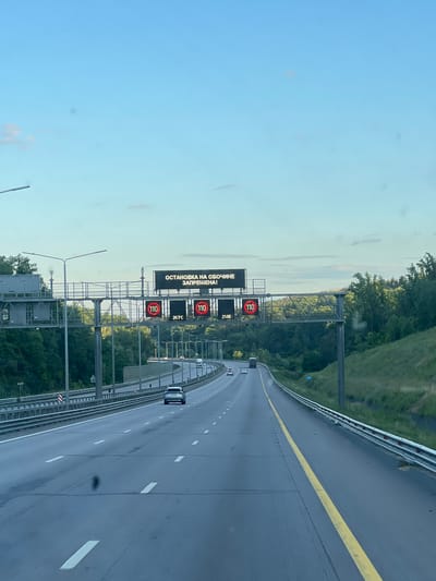 The image captures a highway perspective from a vehicle, looking forward. A digital billboard overhead displays the Russian text "ОСТАНОВКА НА ОБОЧИНЕ ЗАПРЕЩЕНА!" which translates to "Stopping on the shoulder is prohibited!". Below this, multiple digital signs are visible. Two circular signs show "110" in red, indicating a speed limit of 110 km/h. Between these, a sign displays "20.7°C", indicating the temperature. Further along, another "110" sign is present, and a sign shows "2106", likely representing a time or other indicator. The highway itself is paved, with white lane markings, and bordered by a metal guardrail. Lush green trees and foliage line the sides of the road, suggesting a rural or suburban setting outside a city. Several vehicles, including cars and a truck, are driving on the highway. The sky above is a clear blue with a few wispy clouds. There is no visible indication in the image to confirm the location in the city of Voronezh, Russia.