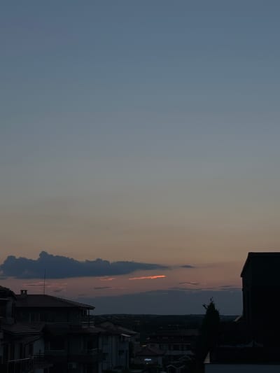 The image depicts a twilight scene over the city of Lozenets, Bulgaria. The sky transitions from a pale blue at the top to a soft orange and grey near the horizon, indicating the sun has recently set. Several dark, textured clouds are visible, some with a faint orange glow from the setting sun. Two small dark shapes, possibly birds, are in flight in the upper part of the sky.
In the foreground and mid-ground, the silhouettes of residential buildings are apparent. These structures appear to be multi-story, with visible rooftops and architectural details like balconies. One building on the right side of the image is a dark, imposing silhouette. The overall impression is of a quiet evening settlement. The environment is urban, characterized by the presence of housing and the fading light of day. There are no identifiable people or ongoing activities.