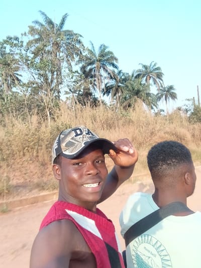 Two young African men pose for a selfie against a bright sky and a landscape featuring tall, dry grasses and palm trees. The man in the foreground wears a camouflage baseball cap, a red and white sleeveless shirt, and smiles. The man behind him wears a light-colored t-shirt with dark straps across his back. The scene suggests a tropical or subtropical environment.