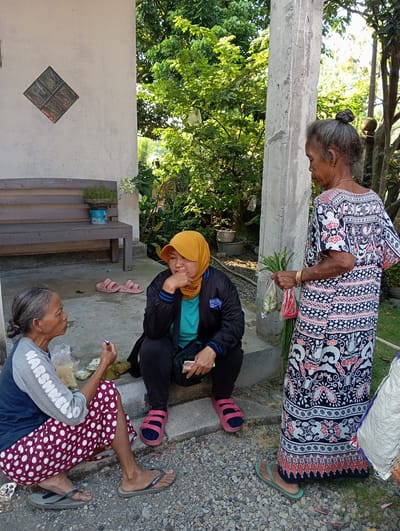 This outdoor image, likely taken in Baron, Indonesia, captures three women engaged in a casual gathering under bright daylight. The scene features concrete ground mixed with gravel, surrounded by lush green foliage and trees, suggesting a warm, tropical climate.
In the upper part of the frame, an older woman stands, wearing a long dress adorned with black, red, and white batik-like patterns. Her grey hair is neatly tied in a bun, and she wears a gold bracelet on her right wrist. She holds a bundle of fresh vegetables, including long beans and red chilies, wrapped in clear plastic. She is wearing green flip-flops.
Below her, two other women are seated on the ground. One woman, appearing middle-aged, wears a black jacket over a blue top, a yellow hijab, and pink sandals. She is resting her chin on her hand, looking downwards with a contemplative or weary expression. Another pair of pink sandals are placed nearby.
To her right, an older woman is seated, wearing a grey long-sleeved shirt that reads "MARSHMELLOW" in white letters across the chest, paired with a dark blue garment and a red and white polka-dotted skirt. She wears black flip-flops and is looking upwards, possibly engaged in conversation or observing the standing woman. A small plastic bag, possibly containing food or other items, is visible near her.
The background features dense green trees and a white wall with a decorative diamond-shaped glass block window. A concrete pillar or beam runs horizontally across the middle of the frame, and a wooden bench with a small potted plant is visible on the left. The overall atmosphere is candid and relaxed, portraying an everyday social interaction.