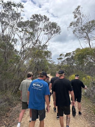 Five men are walking on a dirt path surrounded by trees. They are all wearing casual clothing, including shorts and t-shirts. The man in the front is wearing a blue t-shirt with a Quiksilver logo. The man behind him is wearing a plaid shirt. The others are wearing black shirts. Some are wearing baseball caps. The sky is cloudy.