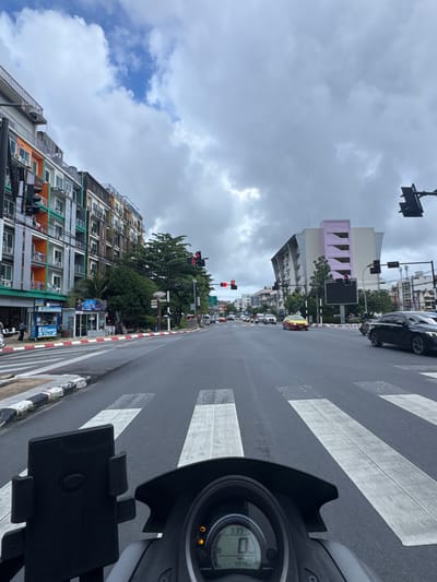 The image depicts a street scene in Patong, Thailand, likely during the daytime given the lighting, under a cloudy sky. The perspective is from the rider of a scooter, looking forward. A zebra crossing is prominently featured in the foreground, with the scooter dashboard visible on the right side, showing "339" and "4948" along with an orange indicator light.
The street is lined with multi-story buildings, some appearing to be apartments or hotels, with balconies and air conditioning units. The architecture features a mix of colors, including orange, green, and gray. One building has a distinctive pastel pink section with arched cutouts. Traffic lights are visible, with one showing a red signal. Vehicles, including cars and possibly other motorcycles, are present in the distance, suggesting ongoing traffic. A yellow taxi is also visible.
The overall scene suggests a typical urban environment with active street life. The cloudy weather might indicate a possibility of rain or just an overcast day. There are several signs visible on the buildings, including "EXCHANGE" signs, indicating commercial activity. A digital billboard is also present on one of the buildings. The image captures a moment of everyday movement within the bustling streets of a Thai tourist destination.