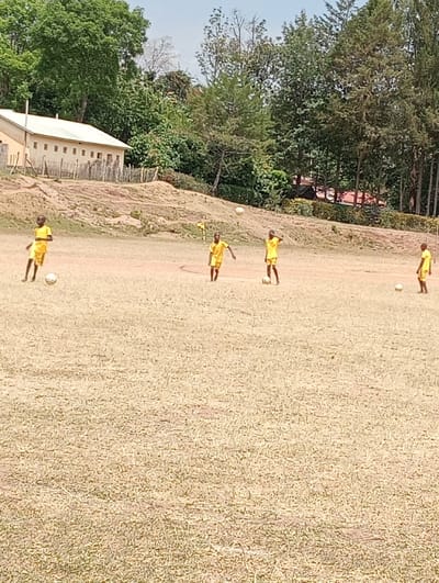 The image shows four young boys playing soccer on a field in Kipkarren, Kenya. The boys are wearing yellow jerseys and shorts. The field is covered in short, dry grass. There are a few trees and a building in the background. The boys are kicking the ball around, having fun.  The boys are playing on a dirt field, likely not a regularly-maintained sports field. The boys are having fun, kicking a soccer ball around the field. The building in the background is a small, single-story structure, perhaps a school or community building. The trees are all deciduous and appear to be in a dry, arid region. There are no other people or objects present. The image captures a scene of leisure and play in a rural area.