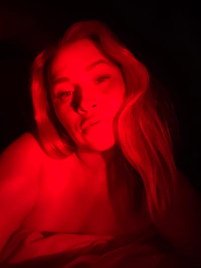 The image shows a close-up of a person's face and upper body, illuminated by a red light against a dark background. The person has long, reddish-blonde hair and is looking towards the camera. The lighting creates strong shadows and highlights on their features, particularly on the forehead, nose, and lips. The upper chest and shoulder area are visible, appearing to be covered by fabric that is also red-lit. The overall impression is moody and intimate. The provided hints focus on different parts of the face, hair, and shoulder, emphasizing the detail and texture captured under the red light. There is no discernible information about the location within Veliky Novgorod, Russia, or any specific objects or actions beyond the visual presentation of the person.