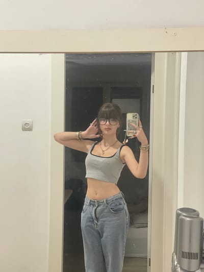 A young woman with dark hair and bangs, wearing glasses, a grey cropped tank top, and light wash jeans, stands in front of a large mirror, taking a selfie. She is adorned with multiple rings on her fingers and bracelets on her wrist. The mirror reflects a room with a dark background. To the left of the woman, a white wall is visible with a single electrical outlet. The scene appears to be indoors, likely a domestic setting, as indicated by the mirror and the interior wall. There is no indication of the city of Ramat Gan, Israel, in the image.