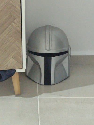 The image presents an indoor scene primarily featuring a silver Mandalorian-style helmet. The helmet, detailed with a distinctive black T-shaped visor and black accents, rests on a light-colored tiled floor. A subtle ridge pattern is visible along the top of the helmet.
To the left of the helmet, a section of furniture is partially visible. This piece features a side panel with a light wooden finish, patterned in what appears to be a herringbone or chevron design. A thin, light-colored border outlines this wooden section. Beneath the furniture, a single, slender wooden leg extends to the floor. A piece of dark blue or black fabric, possibly clothing, is visible near the base of the furniture and its leg.
The floor consists of large, square, light-toned tiles with visible grout lines, suggesting a modern interior. A plain, light grey wall serves as the background behind the helmet. The lighting is diffused and consistent, indicating an indoor setting without strong directional light, offering no clues about the time of day. There are no people, explicit activities, or visible text in the image. The overall atmosphere is quiet and still, depicting objects in a static arrangement within a domestic or display environment, located in Playa del Carmen, Mexico.