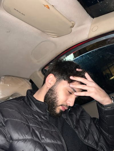 A close-up, candid shot captures a young adult male resting or sleeping inside a car, likely in Joubar Municipality, Syria. The man is seen in profile, with his eyes closed or nearly closed, and his left hand gently placed on his dark, wavy hair. He has a well-groomed dark beard and mustache, and is wearing a black puffer jacket over a dark underlayer. A metallic watch is visible on his left wrist.

The setting is the interior of a vehicle, characterized by light-colored fabric upholstery on the headliner and pillars. To the left, a beige sun visor is visible, showing signs of wear and tear with some reddish-brown marks. Faint, partially obscured text on the visor reads "AIR BAG. See Owner Man...", suggesting "Owner Manual." In the bottom right, a light-colored object, possibly a seat or headrest, is wrapped in clear plastic. The background visible through a window or mirror appears dark, implying low light conditions, such as evening or night. The man's posture and closed eyes convey a sense of repose or weariness.