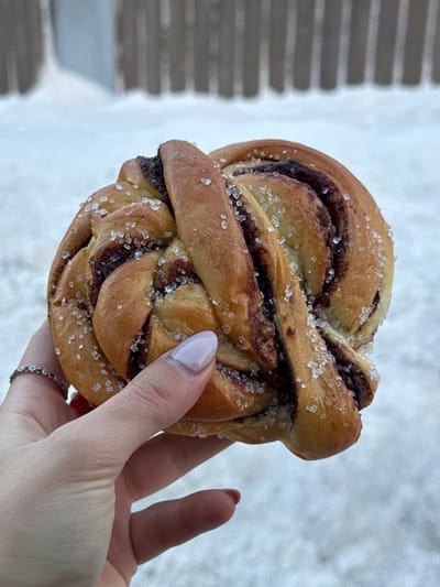 A forensic description of the image shows a hand holding a twisted bun with a dark filling, topped with large sugar crystals, against a snowy outdoor backdrop in Tromsø, Norway. The bun exhibits a golden-brown exterior with visible dark brown filling within its spirals. The hand, with light-toned skin, displays manicured fingernails painted a light lavender hue. A thin, silver-colored ring with small dark beads is worn on the ring finger of the hand. The background is blurred, showing a white, snow-covered ground and a dark, vertically slatted structure, consistent with a winter outdoor setting.