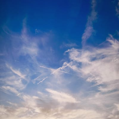 The image is a photograph of a bright blue sky with wispy white clouds. There are several contrails visible, indicating the presence of aircraft. The bottom of the image shows some darker, thicker clouds, possibly at a lower altitude, with a hint of warm light, suggesting it might be near sunrise or sunset. The scene is entirely natural and does not contain any elements that would place it specifically within the city of Knowsley, United Kingdom, or any other city.
