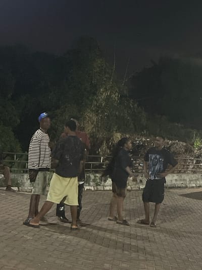 Four individuals stand on a paved walkway, seemingly at night. The person closest to the viewer wears a blue hat, a black and white striped shirt, and dark footwear. Behind them, a person wears yellow shorts, a dark jacket, and dark shoes with white socks. The next figure wears a red shirt and dark jacket, while the furthest individual wears a black shirt with a graphic design and black shorts. A dark, overgrown area is visible in the background, with a railing or wall behind the group.