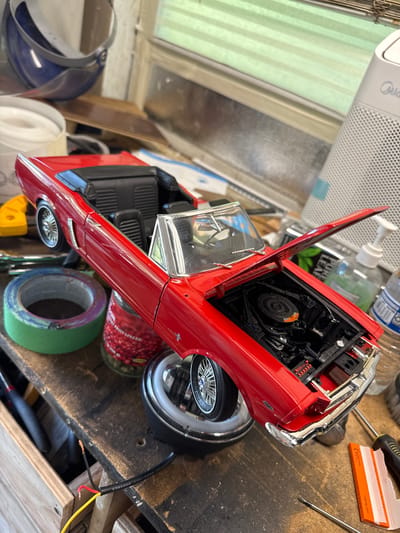 An overhead shot shows a red model convertible with its hood open, revealing a detailed engine. The car is on a wooden workbench, surrounded by various tools and craft supplies, including tape, jars, and bottles. The background suggests an indoor workspace, possibly a garage or workshop, with blinds on a window and a white electronic device. The overall scene depicts a model car assembly or hobbyist setup in North Highlands, United States.