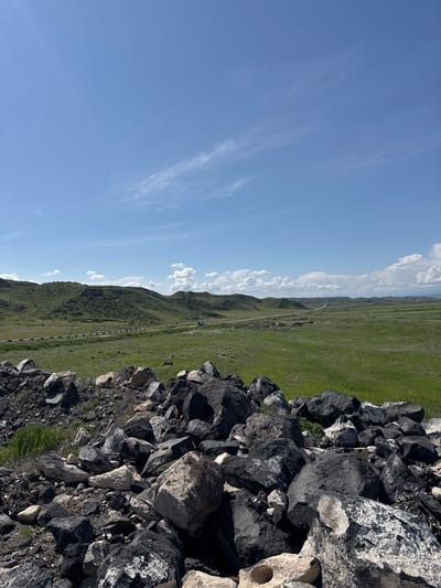 The image shows a landscape in Aniavan, Armenia, featuring a rock wall or pile in the foreground. Beyond the rocks is a green, grassy field with a road or path running through it. In the distance, there is a horizon line marked by hills and clouds. The sky is blue with scattered clouds. There are no people visible.