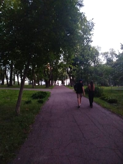 The image shows a paved pathway in a park setting, lined with trees. Two individuals are walking away from the camera down the path. The person on the left is wearing a dark t-shirt, light-colored shorts, and a blue cap, carrying a dark backpack. The person on the right is wearing dark clothing. The pathway appears to be made of asphalt and shows some cracking. To the left of the path is a grassy area with a tree trunk in the foreground and a dense line of trees further back. To the right, there is also a grassy area with vegetation. The lighting suggests it is either early morning or late evening, with a diffused light source from the sky. The park appears to be a public space. Without further contextual information, the specific location within Chernihiv, Ukraine, cannot be determined from the image alone.