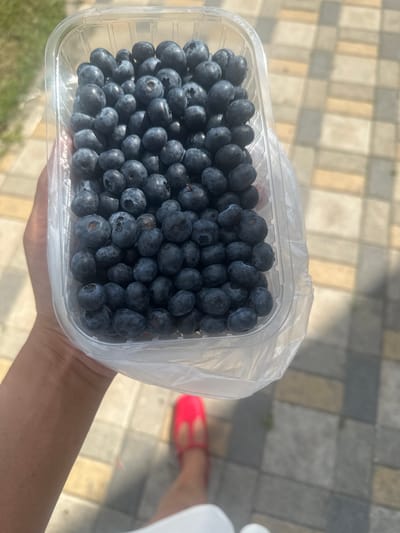 A hand holds a clear plastic container filled with blueberries. The container is partially wrapped in plastic film. The background shows a paved area with rectangular tiles in shades of beige and gray. A person wearing bright pink open-toed shoes is visible at the bottom of the frame. The scene is outdoors, likely on a sunny day, given the lighting. The location is not identifiable as Moskva, Russia.