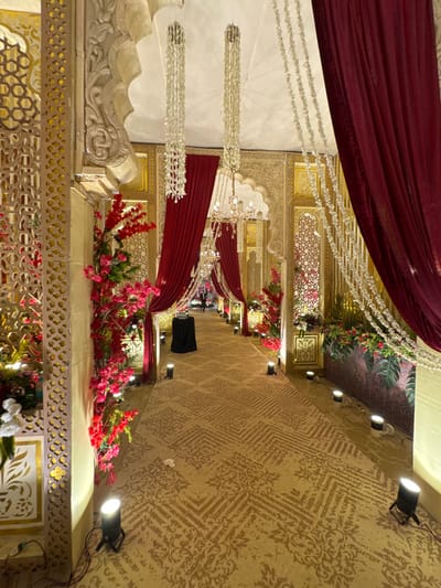 A decorative indoor space showcases a luxurious setup with burgundy drapes and ornate gold accents. The walls feature patterned wallpaper, while the ceiling is adorned with floral arrangements and hanging crystal strands. Red and pink flowers are strategically placed along the perimeter. Modern spotlights illuminate the scene, creating a warm and inviting ambiance.