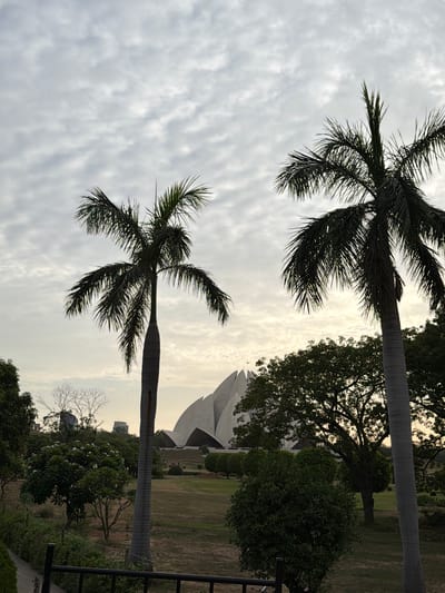 The image depicts an outdoor scene with two palm trees prominently featured in the foreground. The palm trees are silhouetted against a cloudy sky, suggesting it might be a partly cloudy or overcast day. In the background, a modern architectural structure with large, petal-like white surfaces is partially visible, set within a landscape of green grass, trees, and bushes. This structure resembles the Lotus Temple in Delhi, India, a significant landmark known for its flower-like design and used as a place of worship for people of all faiths. The time of day appears to be late afternoon or early evening, as indicated by the soft, diffused light. The overall atmosphere is serene and tranquil, with no people visible in the frame. The presence of palm trees and the architectural style of the background strongly suggest the location is Delhi, India. There is no visible text in the image.