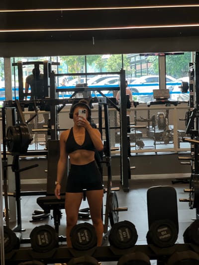 The image shows a young woman taking a selfie in a gym. She is wearing a black sports bra and black shorts, with headphones on. She is standing in front of a mirror, with gym equipment visible in the background, including weight racks, dumbbells, and weight machines. Through the large windows in the background, a street scene with cars and trees can be seen. The location is identified as Si Sunthon, Thailand, suggesting this is a gym in that city. The lighting in the gym appears to be artificial, with overhead fluorescent lights. The overall atmosphere is one of a typical fitness center.