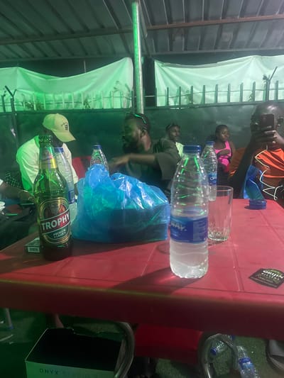 The image shows a gathering of people around a red table. On the table are multiple water bottles, a bottle of Trophy beer, and a drinking glass. The location is Karu, Nigeria, based on the beer brand "Trophy Lager." The scene appears to be outdoors, with a makeshift tent or awning visible. People are sitting and standing around the table, engaged in conversation and socializing. One person is holding a mobile phone. An Onyx Studio speaker is on the ground.