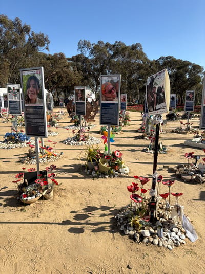 The image displays an outdoor memorial site, specifically the Nova Festival Memorial, located in the Eshkol Regional Council, Israel. Numerous vertical memorial stands are arranged in rows across a light brown, sandy ground. Each stand consists of a grey metal pole supporting a rectangular display board that features a prominent color photograph of an individual, typically a young person, with descriptive text primarily in Hebrew below it, and often a heart icon at the top. The base of each stand is surrounded by a circular arrangement of small, light-colored pebbles. Placed within or upon these pebbles are various commemorative items, including numerous artificial red flowers resembling poppies on wire stems, small potted green plants, candles, pinwheels, small flags (e.g., Israeli, Canadian), and other personal mementos. One stand includes a large, dark brown, abstract metal sculpture of an animal. Another stand incorporates a white, curved object behind its display board. The sandy ground exhibits numerous footprints and scuff marks. The background features a dense line of tall trees with dark green foliage under a clear, cloudless blue sky, with strong sunlight casting distinct shadows from the stands and flowers.