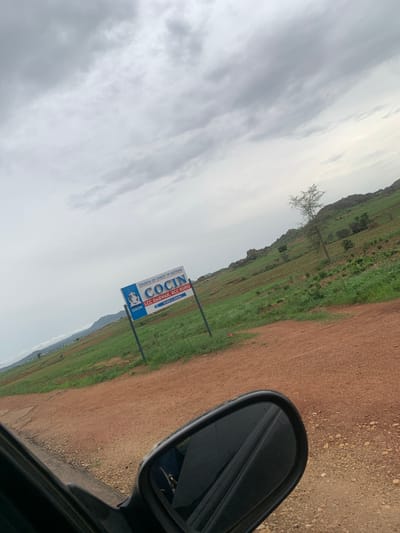 The image shows a sign for "CHURCH OF CHRIST IN NATIONS COClN LCC DABWAK, RCC KURU PO. BOX 12, KURU" in Kuru Dungus, Nigeria. The sign is blue and white and features a logo with a dove. The sign is set against a backdrop of a green, hilly landscape under a cloudy sky. A dirt road is visible in the foreground, and the side mirror of a vehicle can be seen on the right. The location appears to be rural, with agricultural fields and scattered trees.