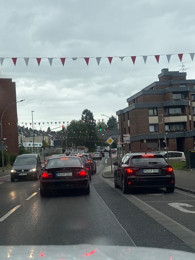 The image captures an urban street scene in Mönchengladbach, Germany, identifiable by the "MG" prefix on vehicle license plates (MG XX 2016, MG LR 1010, MG HK 2912) and a visible street sign for "Untere Straße." The weather is overcast with a cloudy sky, and the asphalt road surface is wet, reflecting light, suggesting recent rain.
Red and white triangular bunting is strung across the street overhead, spanning from left to right. In the immediate foreground, a dark red BMW 3-series sedan, license plate MG XX 2016, is centered, its brake lights illuminated as it queues in traffic. To its right, a dark gray Audi A1 Sportback, license plate MG LR 1010, is parked partially on a paved curb, also displaying illuminated rear lights. Further ahead in the left lane, a dark gray Volkswagen Transporter van, license plate MG HK 2912, proceeds with its headlights on. Additional vehicles are visible in the traffic queue and parked along the street.
On the left side of the street, a large brick building with a smooth facade is partially visible. In the midground, a traffic light at an intersection displays red, while another further to the right shows green. Road signs include a yellow diamond sign with "Untere Straße" and a circular "30" km/h speed limit sign. On the right side, a multi-story brick apartment building with distinctive tiered balconies and a flat roof dominates the midground. The background features more residential buildings with pitched roofs and mature trees under the consistent cloud cover. No pedestrians are clearly discernible in the scene.